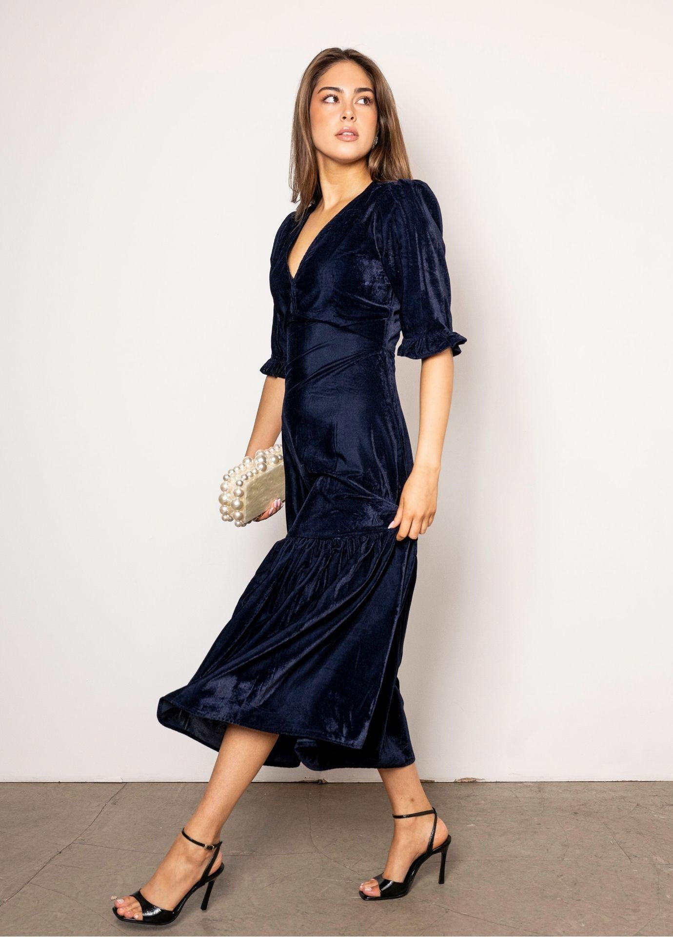 Ophelia Dress Velvet Blue Sapphire: a woman walks in a navy velvet midi dress with a V-neckline, short puff sleeves and a flounced hem, holding a pearl clutch, with black strappy heels on a concrete floor.