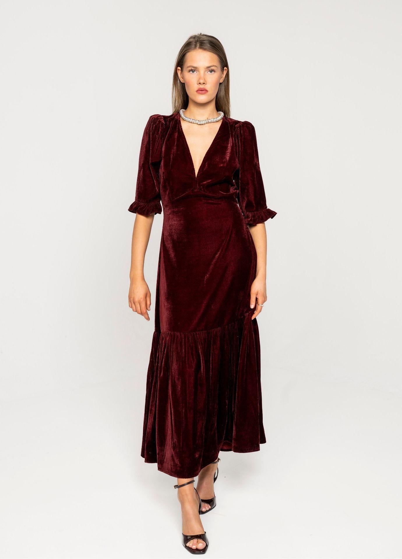 Ophelia Dress Velvet Bordeaux: a woman wears a deep red velvet midi dress with a V-neckline, short puff sleeves with ruffled cuffs and a flounced hem, standing in a light grey studio in black strappy heels.