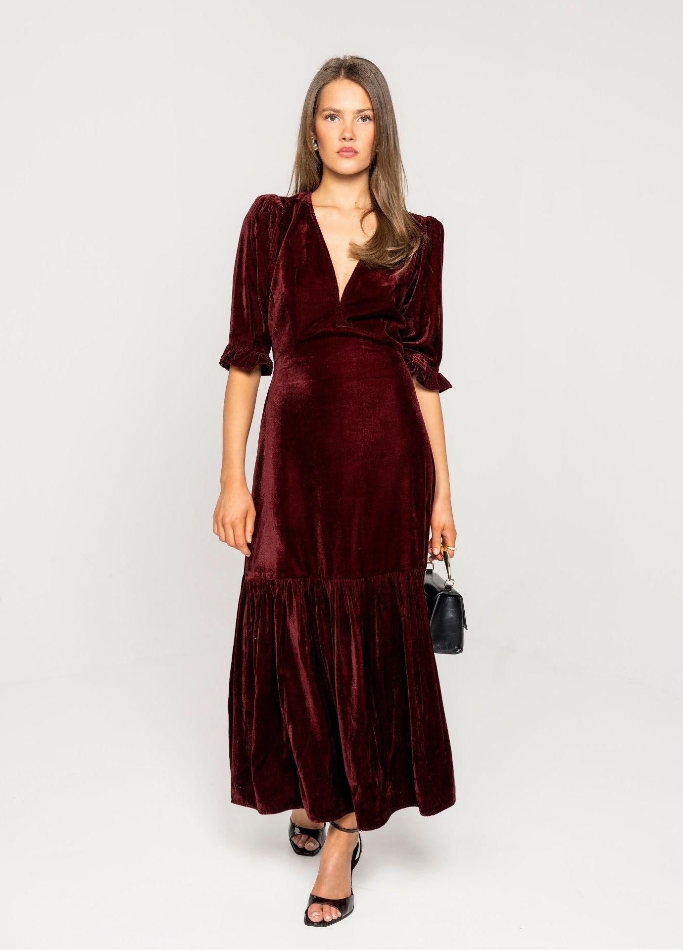 Ophelia Dress Velvet Bordeaux in a full length front view, holding a small black handbag, showing the fitted bodice and the flounced hem.