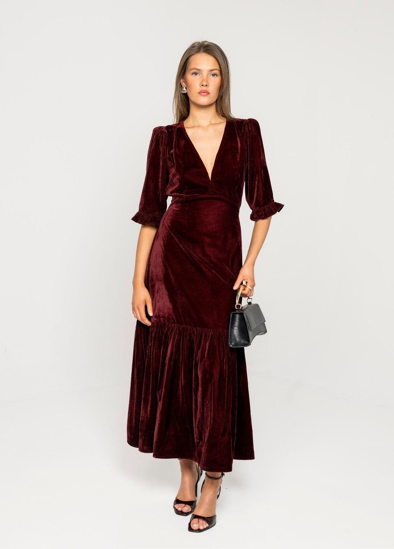Ophelia Dress Velvet Bordeaux in a front view with a small black bag, the red velvet midi dress falling to mid calf against a light grey background.