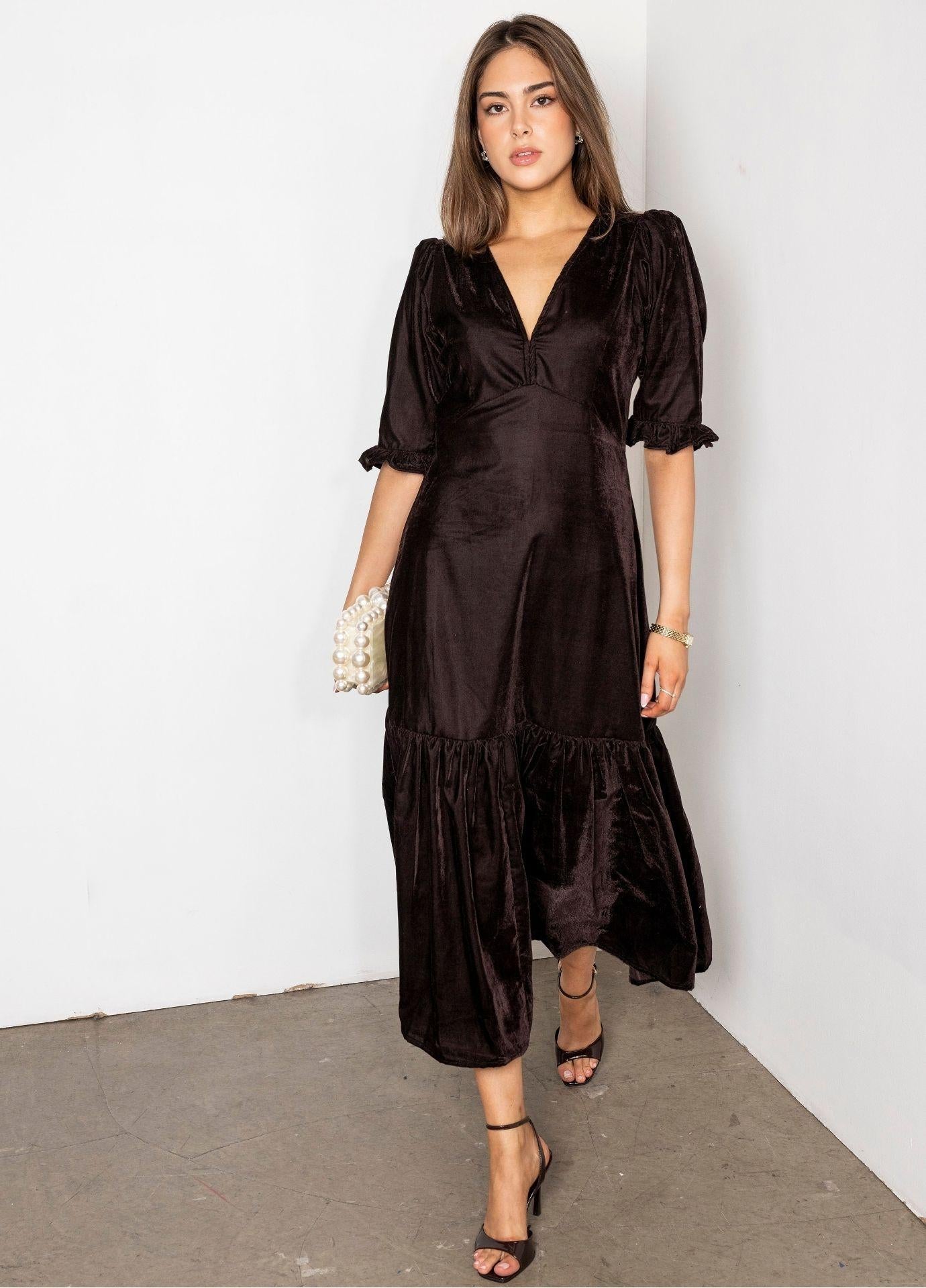Ophelia Dress Velvet Toffee: a woman wears a toffee brown velvet midi dress with a V-neckline, short puff sleeves with ruffled cuffs and a flounced hem, holding a pearl clutch in a white studio corner.