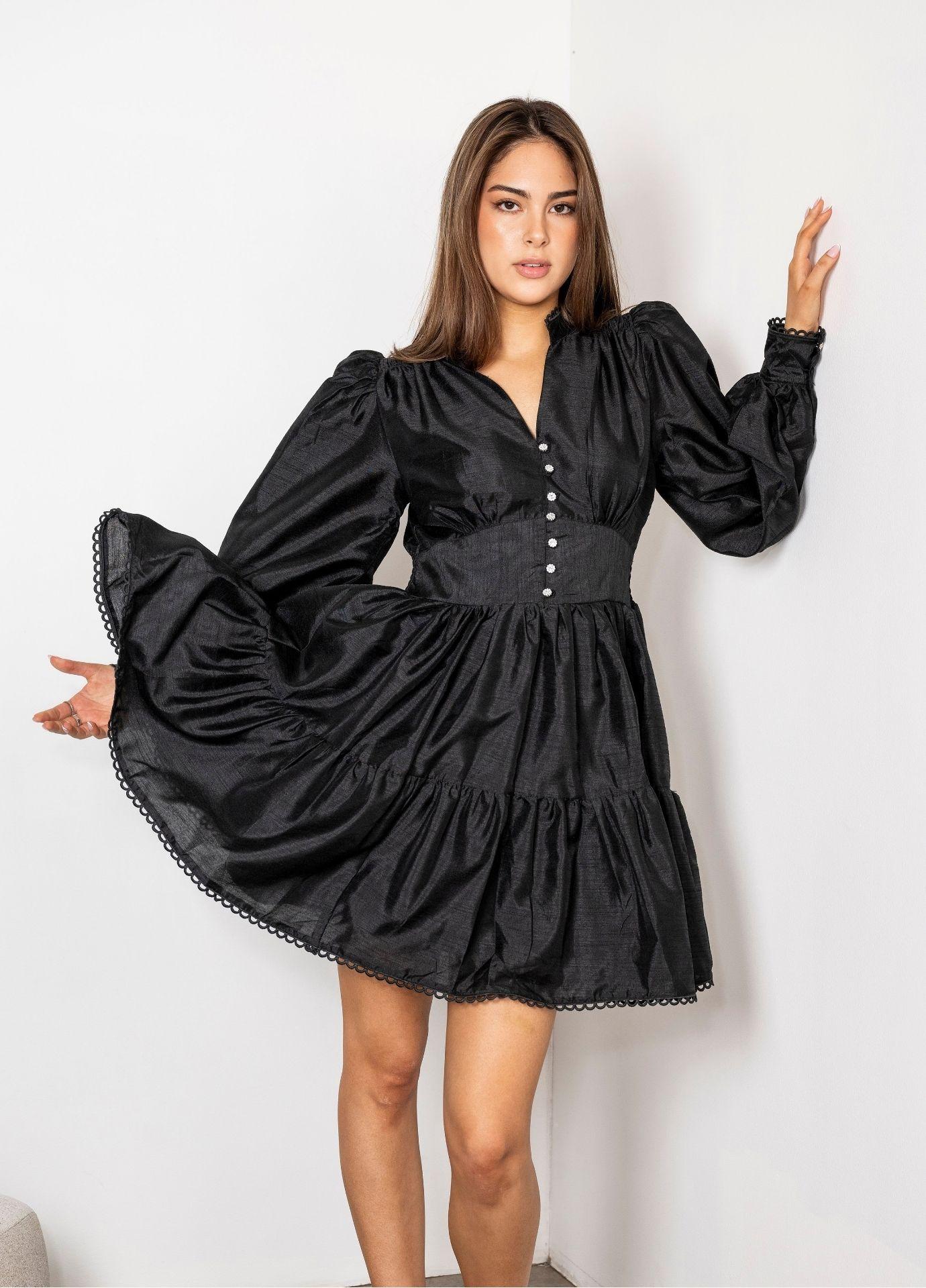 Primrose Dress Evening Black: a woman twirls in a black mini dress with a soft sheen, voluminous balloon sleeves, a wide waistband and a tiered skirt trimmed with lace, against a white wall.