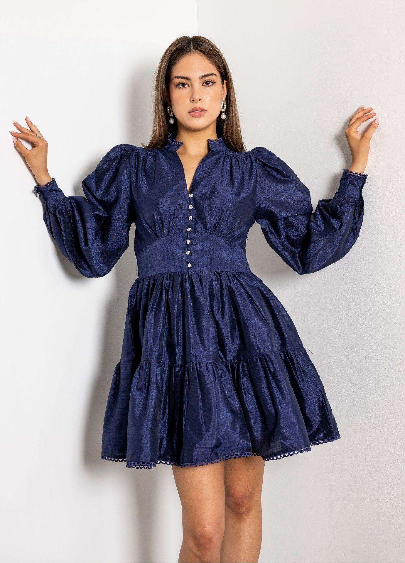 Primrose Dress Evening Blue: a woman raises her arms against a white wall, wearing a navy mini dress with voluminous balloon sleeves, a high collar with a V opening and small pearly buttons down the bodice.