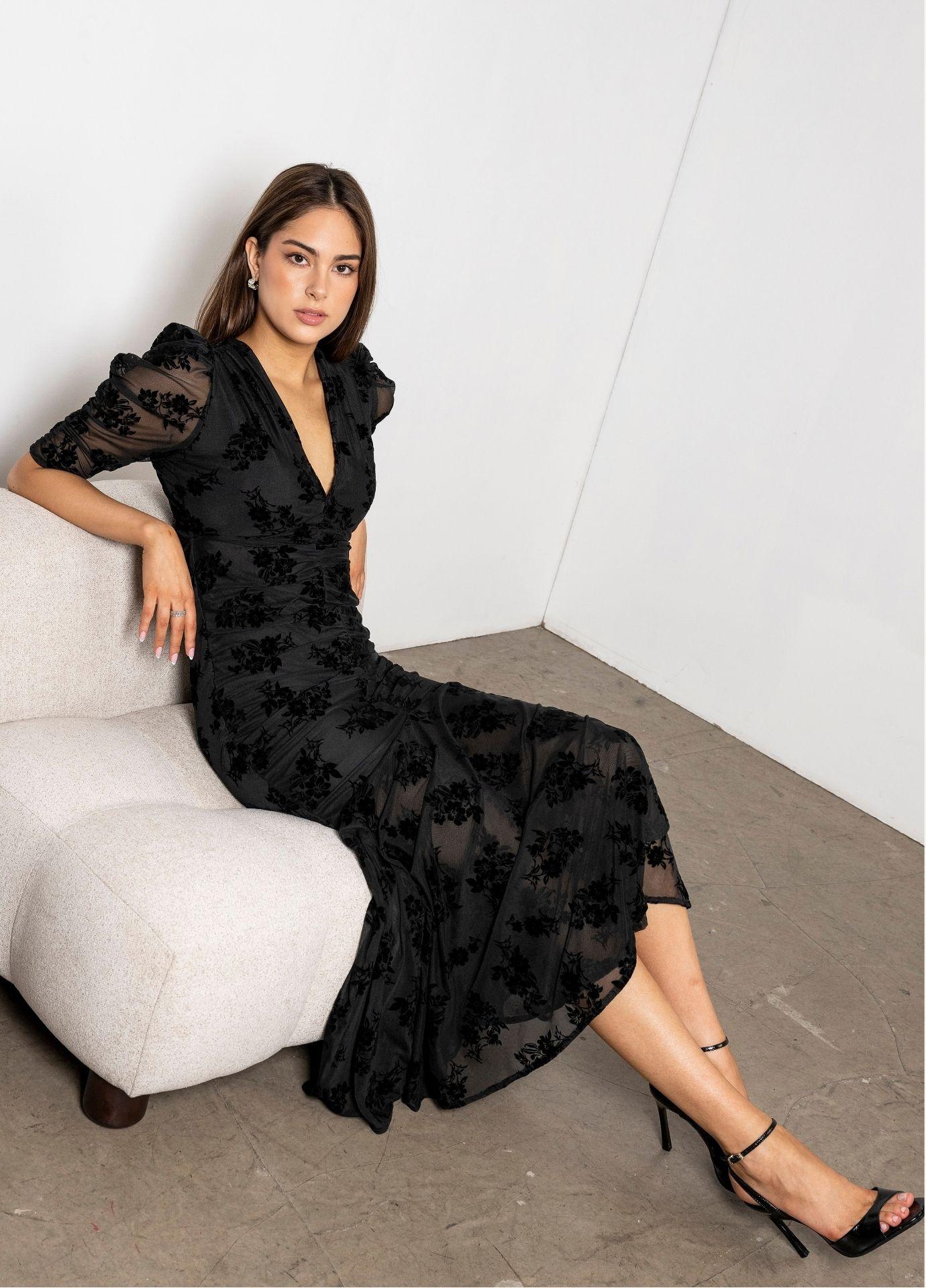 Sofia Dress Blooming Mesh: a woman sits on a cream upholstered chair wearing a black mesh maxi dress with velvety flowers, a V-neckline and ruched puff sleeves, with black strappy heels on a concrete floor.