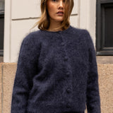A woman wearing a fuzzy dark navy blue button-front cardigan sweater, standing outdoors against a light-colored building wall.