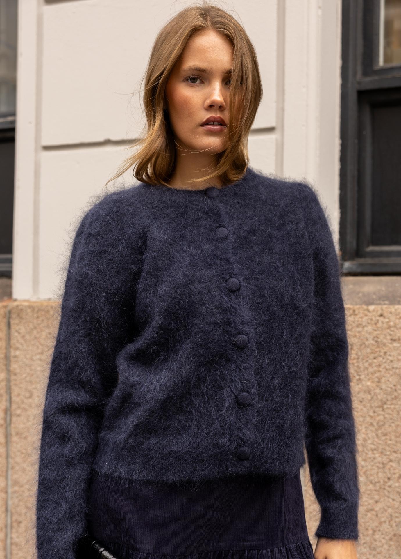 A woman wearing a fuzzy dark navy blue button-front cardigan sweater, standing outdoors against a light-colored building wall.