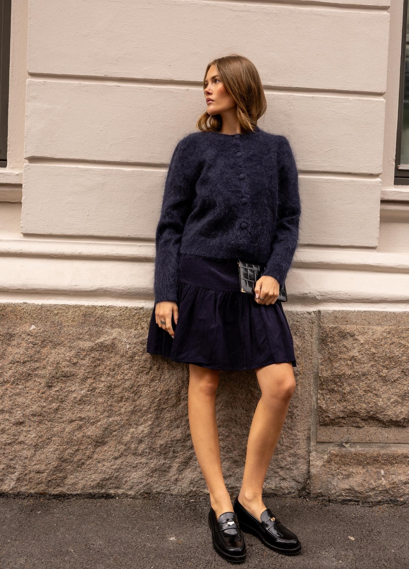 A woman wearing a fuzzy dark navy blue button-front cardigan sweater, standing outdoors against a light-colored building wall.