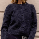 A woman wearing a fuzzy dark navy blue button-front cardigan sweater, standing outdoors against a light-colored building wall.