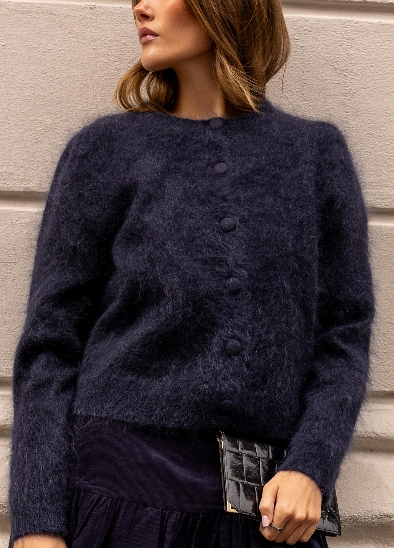 A woman wearing a fuzzy dark navy blue button-front cardigan sweater, standing outdoors against a light-colored building wall.