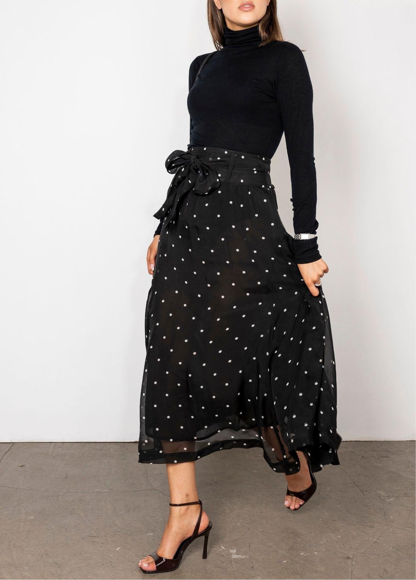 Vanja Skirt Black Pearl: a woman wears a sheer black midi skirt scattered with small white dots, tied with a bow at the waist and styled with a black turtleneck, in a full length side view.