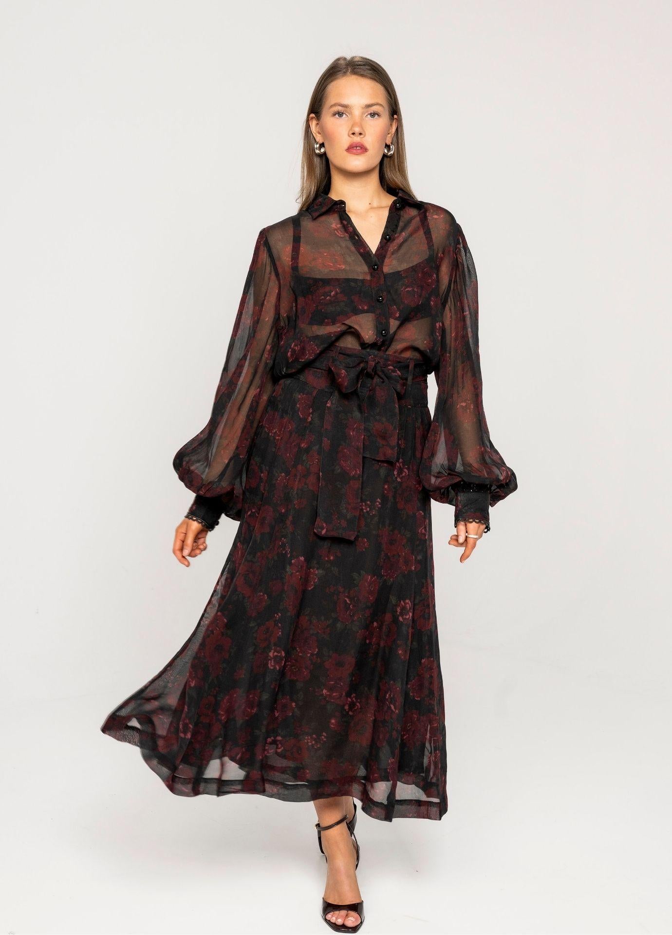 Vanja Skirt Black Romance: a woman walks in a sheer black midi skirt with a deep red floral print, worn with the matching Anaya Blouse Black Romance and black strappy heels in a light grey studio.