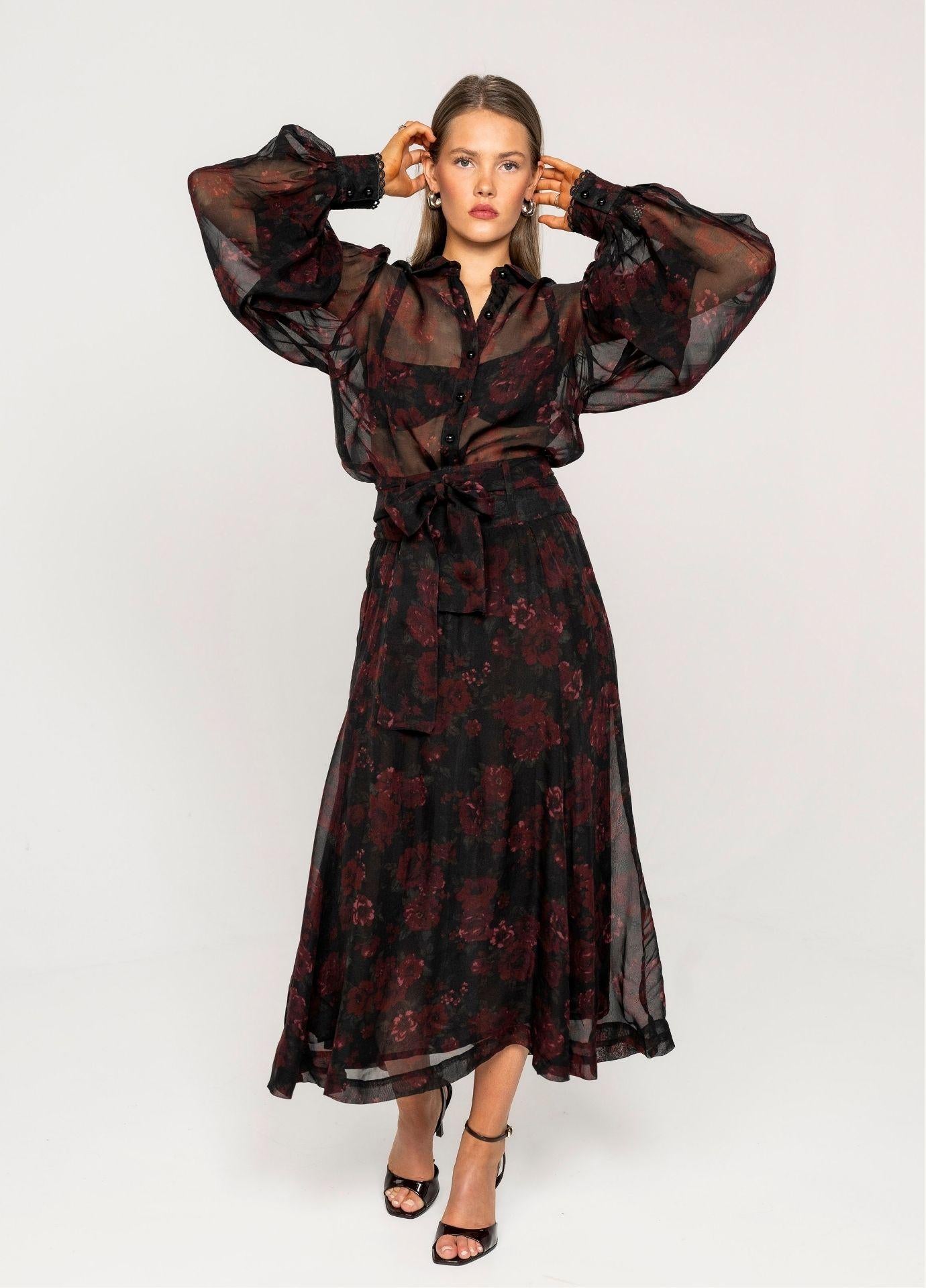 Vanja Skirt Black Romance with the model's hands raised to her hair, styled with the matching sheer floral blouse with balloon sleeves against a light grey background.